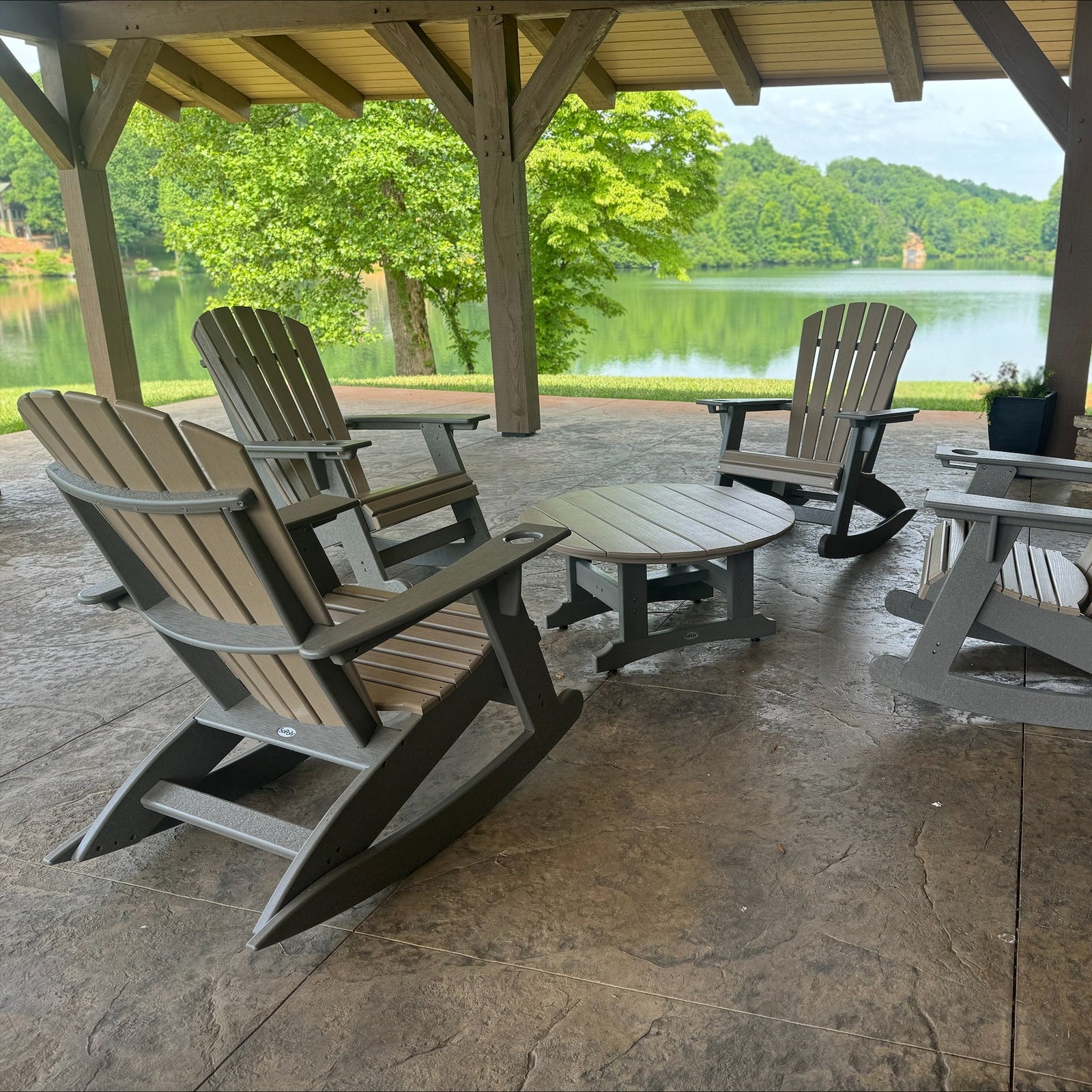 Four gray Adirondack-style outdoor chairs arranged around a small round table on a covered patio, with a stone fireplace and a lake view in the background.